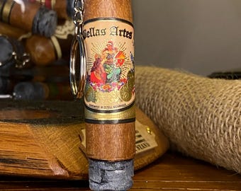 May include: A cigar-shaped keychain with a gold-colored ring and chain. The cigar has a brown body with a label that reads "Bellas Artes" and features a colorful illustration. The end of the cigar is gray and black.