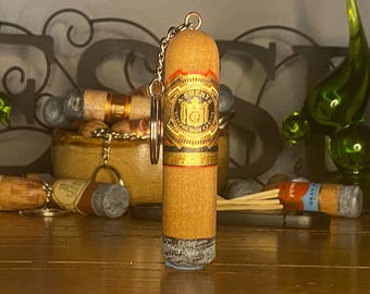 May include: A cylindrical cigar-shaped keychain featuring a light brown wooden body with a detailed Arturo Fuente Reserva Don Carlos cigar band in red, gold, and black. The base of the keychain has a textured grey ash-like finish and a metal ring.