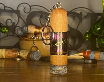 May include: A cylindrical wooden keychain shaped like a cigar, featuring a metallic band with the text ACID printed in gold. The bottom end has a textured gray finish resembling ash, and a silver metal split ring is attached to the top.