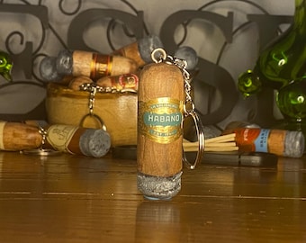 May include: A brown cigar-shaped keychain featuring a gold and teal band with the text HABANO. The bottom of the cigar has a textured grey ash effect. A silver metal chain and ring are attached to the top of the cigar-shaped object.