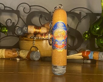 May include: A cylindrical wooden keychain shaped like a cigar, featuring a blue and gold label with the text San Lotancy printed in the center. The object has a grey textured tip at the bottom and a metal ring attached to the top for hanging.