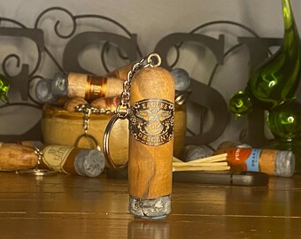 May include: A cylindrical wooden keychain shaped like a cigar, featuring a black band with the text Eagles Descent. The bottom end has a textured gray ash-like finish, and a silver metal chain and ring are attached to the top of the wooden body.