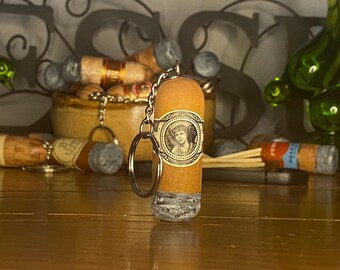 May include: A cylindrical cigar-shaped keychain made of light brown wood with a textured gray ash tip. The center features a decorative paper band with a circular illustration of a person's face surrounded by ornate gold and black border details.