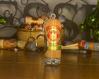 May include: A cylindrical keychain shaped like a cigar with a textured gray ash tip. The body features a red, green, and gold band with the text A. FUENTE GRAN RESERVA printed in white and gold. A silver metal chain and ring are attached to the top.