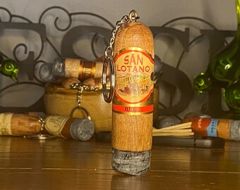 May include: A cylindrical wooden keychain shaped like a cigar, featuring a prominent red and gold label with the text SAN LOTANO BULL. The top includes a metal chain and ring, while the bottom end has a textured, grey, ash-like finish.