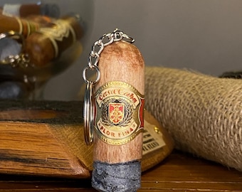 May include: A cigar-shaped keychain with a gold-colored label that reads "Casa Cuba". The keychain has a silver chain and ring. The cigar has a light brown body and a dark gray base.