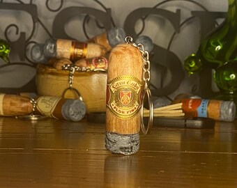 May include: A cigar-shaped keychain featuring a light brown wooden body with a textured gray ash tip. A gold and red band is wrapped around the center, displaying the text Casa Cuba Flor Fina with a central crest. A silver metal chain is attached.