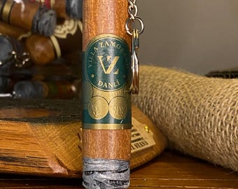 May include: A wooden cigar-shaped keychain with a silver chain and key ring. The cigar has a green and gold label that reads "Villa Zamorano Danli". The end of the cigar is wrapped in a silver material.