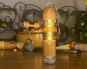 May include: A cylindrical wooden keychain shaped like a cigar with a textured gray ash tip. The body features a prominent yellow, gold, and blue label with the text Diamond Crown Maximus printed in black and gold lettering, attached to a metal ring.
