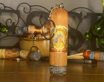 May include: A wooden cigar-shaped keychain featuring a metallic ring at the top. The body displays a decorative label with the text La Aroma de Cuba, an illustration of a person, and the words Handmade Imported. The base has a textured gray finish.