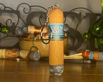 May include: A cylindrical cigar-shaped keychain featuring a light brown textured body with a blue band printed with the text NICA RUSTICA ADOBE. The bottom end has a grey ash-like finish, and a silver metal chain and ring are attached to the top.