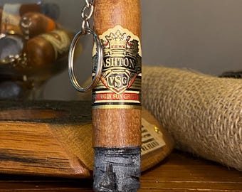 May include: A cigar-shaped keychain with a metal ring and chain. The cigar has a brown wooden body with a label that reads "Ashton VSG". The bottom of the cigar is dark gray and blue.