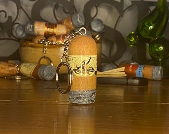 May include: A wooden cigar-shaped keychain featuring a tan label with a black graphic of a person and the text "CIGAR RIGHTS OF AMERICA". The bottom of the keychain has a textured gray finish resembling ash, and it includes a silver metal ring.