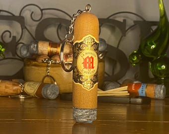 May include: A cylindrical keychain shaped like a cigar with a tan body and a grey textured end. The center features a decorative label with black and gold filigree, a red logo, and the text "LA MISSION L'ATELIER" with a metal ring attached.