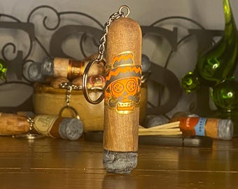 May include: A cylindrical wooden keychain shaped like a cigar with a grey textured end. The surface features a printed orange and black graphic of a skull wearing a sombrero and a mustache. A metal chain and ring are attached to the top of the object.