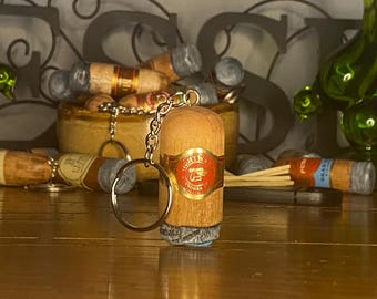 May include: A cylindrical wooden keychain shaped like a cigar, featuring a red and gold band with the text "LIGHTER" and "INORSA" printed on it. The keychain has a silver metal chain and a circular ring attached to the top of the wooden cigar body.