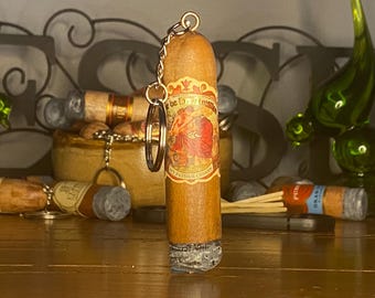 May include: A brown cigar-shaped keychain featuring a decorative paper band with a red and gold illustration of figures and the text My Father Cigars. The bottom end of the cigar has a textured gray ash appearance, and a silver metal ring is attached.