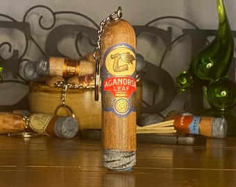 May include: A cylindrical wooden keychain shaped like a cigar, featuring a paper band with the text Aganorsa Leaf Nicaragua and a graphic of an arm holding a bundle of leaves. The bottom end has a textured gray finish resembling cigar ash.