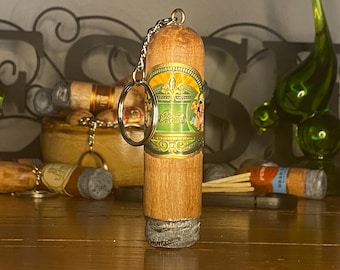 May include: A cylindrical wooden keychain designed to resemble a cigar, featuring a detailed green and gold paper band with ornate patterns and text. The top includes a metal chain and ring, while the bottom end has a textured gray finish.