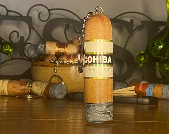 May include: A wooden cigar-shaped keychain featuring a band with the text COHIBA CONNECTICUT in black and red lettering. The bottom of the cigar has a textured gray ash-like finish, and a silver metal chain and ring are attached to the top.