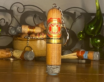 May include: A cylindrical cigar-shaped keychain made of light brown wood with a realistic gray ash tip. The center features a red, gold, and black label with the text A. FUENTE GRAN RESERVA printed in white and gold. A silver metal chain is attached.
