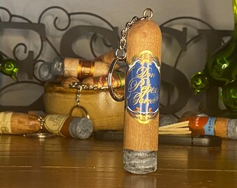 May include: A cylindrical cigar-shaped keychain with a light brown textured surface and a blue and gold label featuring the text Don Pepin Garcia. The bottom end has a grey ash-like finish, and a silver metal chain is attached to the top.