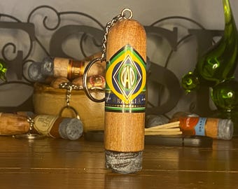 May include: A cylindrical wooden keychain designed to resemble a cigar, featuring a band with the text CAO BRAZILIA in yellow, green, and blue colors. The bottom end has a textured gray finish, and a metal chain and ring are attached to the top.