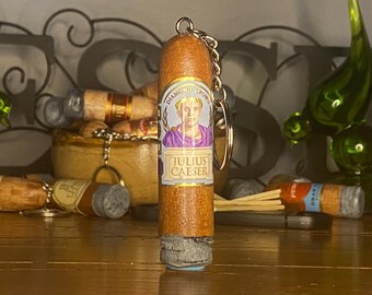 May include: A cylindrical wooden keychain shaped like a cigar with a textured gray end. The label features a portrait of Julius Caesar in a purple toga with a gold laurel wreath, the text "DIAMOND CROWN" and "JULIUS CAESER" printed on it.