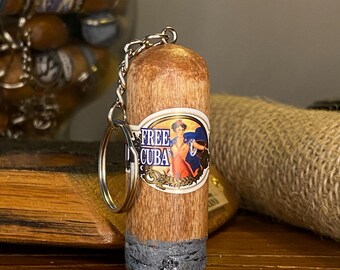 May include: A cigar-shaped keychain with a silver keyring. The keychain has a brown body with a gray base and a label that reads "FREE CUBA" with a colorful illustration. The keychain is made of wood.