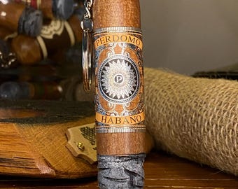 May include: A cigar-shaped keychain with a silver chain and ring. The cigar has a brown body with a decorative band featuring the text "PERDOMO HABANO" and a circular design. The end of the cigar is gray and blue.