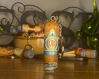 May include: A brown cigar-shaped keychain featuring a blue and gold band with the text CAO NICARAGUA printed on it. The keychain has a silver metal ring attached to the top, and the bottom end of the cigar is textured to resemble gray ash.