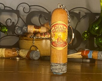 May include: A cylindrical wooden keychain shaped like a cigar, featuring a gold and red label with the text Días de Gloria Nicaragua. The base of the keychain has a textured, grey, bark-like appearance, and a metal chain and ring are attached to the top.