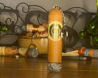 May include: A cylindrical cigar-shaped keychain featuring a light brown wooden body with a Macanudo brand cigar band in the center. The bottom end has a textured grey ash appearance, and a silver metal chain and ring are attached to the top end.