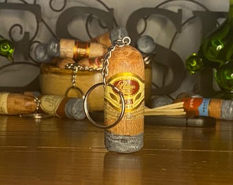 May include: A cylindrical keychain shaped like a cigar with a brown textured body and a grey ash-like base. It features a gold and white label with the text Padrón 1926 Serie and a silver metal ring attached to the top by a small chain.