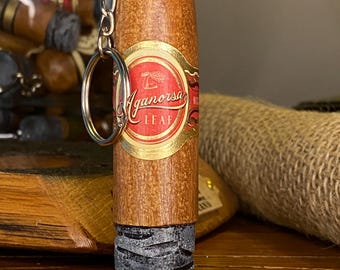 May include: A cigar-shaped keychain with a gold-colored ring and chain. The keychain has a brown body with a red and gold label that reads "Aganorsa Leaf". The bottom of the keychain is dark gray.