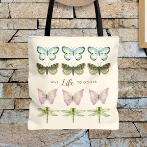 Vintage Butterfly AOP ToteBag! 3 times 3 Butterflies in a row in colours yellow-blue, green, yellow-pink, and 1 time dragonflies in light green. In the middle is a saying: Give live to others.
Tote Bag is hanging on a stone wall, with a summer hat.
