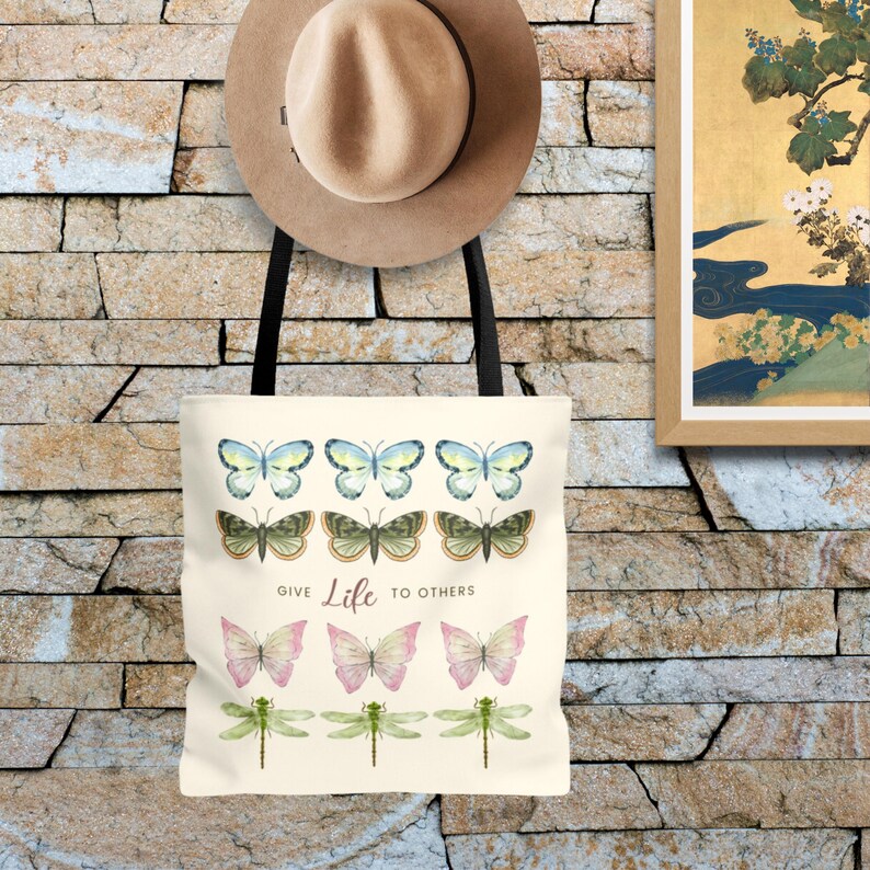 Vintage Butterfly AOP ToteBag! 3 times 3 Butterflies in a row in colours yellow-blue, green, yellow-pink, and 1 time dragonflies in light green. In the middle is a saying: Give live to others.
Tote Bag is hanging on a stone wall, with a summer hat.