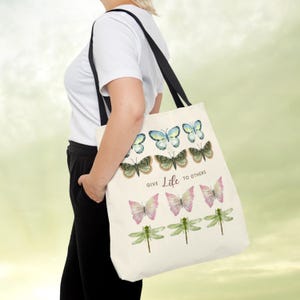 Vintage Butterfly AOP ToteBag! A woman has a Tote Bag on her shoulder. The tote bag has 3 times 3 Butterflies in a row in colours yellow-blue, green, yellow-pink, and 1 time dragonflies in light green. In the middle is a saying: Give live to others.