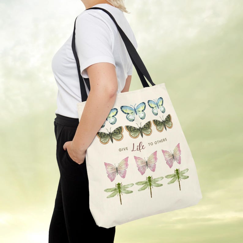 Vintage Butterfly AOP ToteBag! A woman has a Tote Bag on her shoulder. The tote bag has 3 times 3 Butterflies in a row in colours yellow-blue, green, yellow-pink, and 1 time dragonflies in light green. In the middle is a saying: Give live to others.