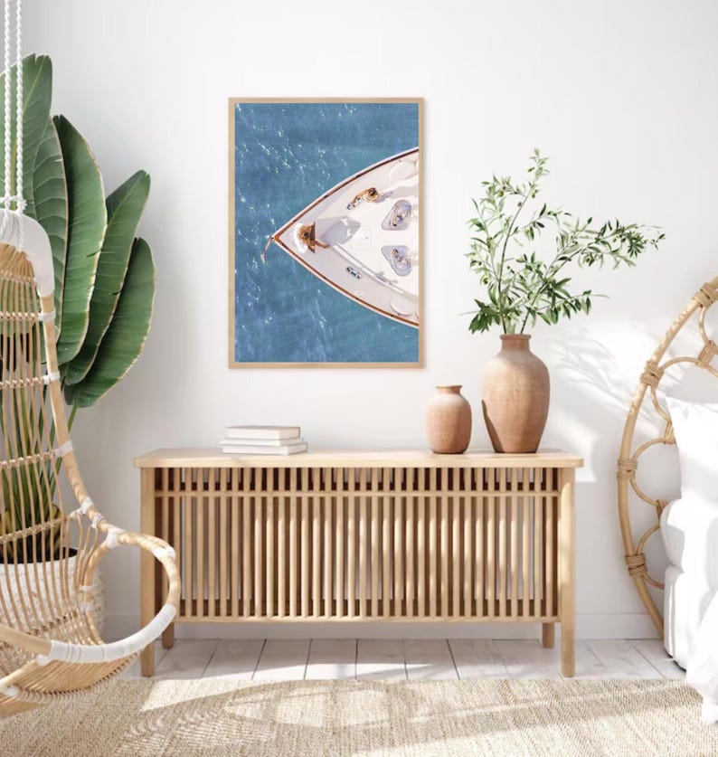 May include: A framed photograph of a white boat on the water. The boat is viewed from above, and the bow of the boat is facing the viewer. The water is a light blue color, and the sky is a light blue color. The photograph is hanging on a white wall.