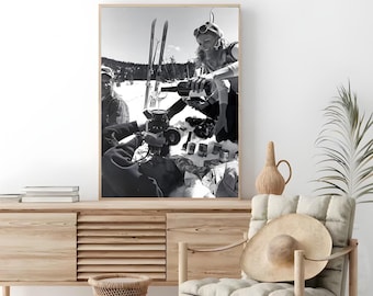 May include: A black and white photograph of a group of people enjoying a picnic in the snow. They are sitting on a blanket and drinking wine from a bottle. There are ski poles and a camera in the background.