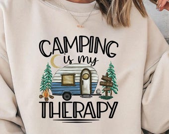 May include: Beige sweatshirt with the text "CAMPING is my THERAPY" in black. The graphic features a blue and white camper, trees, and a campfire. The sweatshirt is a neutral color and has long sleeves.