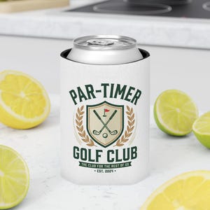 以下が含まれることがあります： 緑と茶色で「PAR-TIMER GOLF CLUB」の文字が入った白いゴルフ缶クーラー。デザインには、交差したゴルフクラブ、ゴルフボール、赤い旗が含まれています。クーラーは、カットされたレモンとライムに囲まれています。