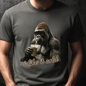 May include: A charcoal gray crew neck t-shirt featuring a graphic of a gorilla wearing black-rimmed glasses and drinking from a clear plastic cup with a straw. Below the gorilla, the text "i like it cold" is printed in a brown cursive font.