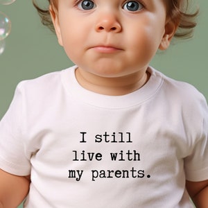 May include: A baby wearing a white onesie with the text "I still live with my parents." printed on it. The baby is looking at the camera with a serious expression.