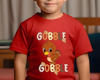 Puede incluir: Una camiseta roja con la palabra "GOBBLE" en una fuente decorativa encima y debajo de un pavo de dibujos animados. El pavo es marrón con un pico amarillo y está rodeado de hojas de otoño. La camiseta la lleva un niño pequeño.