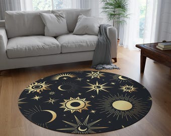 May include: A round black rug featuring a celestial design with gold suns, stars, and crescent moons. The rug is placed on a hardwood floor in a living room setting, with a white sofa and natural light.