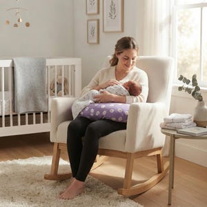 May include: A purple nursing pillow featuring a white daisy pattern, positioned on the lap of a person sitting in a beige upholstered rocking chair with light wooden rockers, while supporting a swaddled infant in a white patterned blanket.
