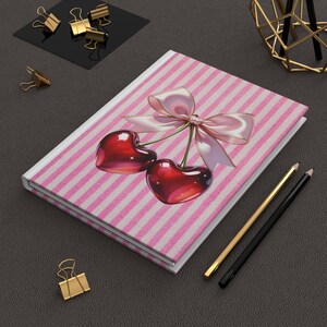 Könnte beinhalten: Ein Notizbuch mit rosa und weißen Streifen, das eine Kirschenillustration und eine rosa Schleife zeigt. Goldene Büroklammern, Bleistifte und ein goldenes geometrisches Objekt umgeben das Notizbuch.