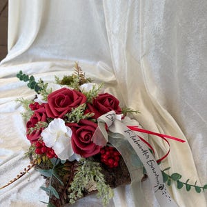 Könnte beinhalten: Ein Blumenarrangement mit dunkelroten und weißen Rosen, roten Beeren und grünem Blattwerk auf einem Stück natürlicher Baumrinde. Ein helles Band ist über das Arrangement drapiert und trägt den Text In liebevoller Erinnerung.